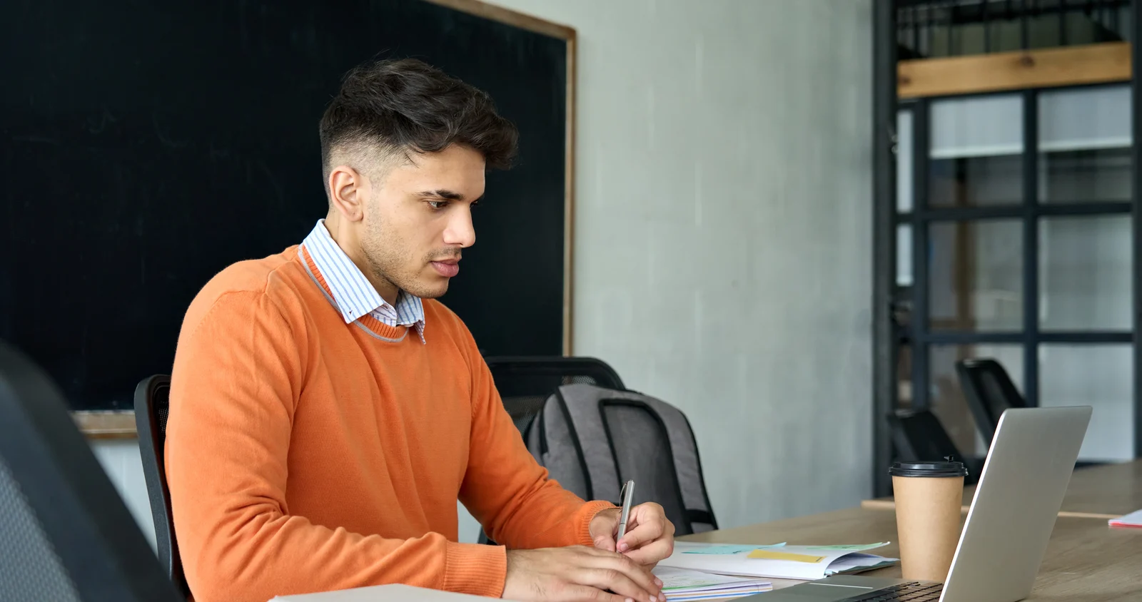 A young man in an orange sweater taking notes beside a laptop at a long study table